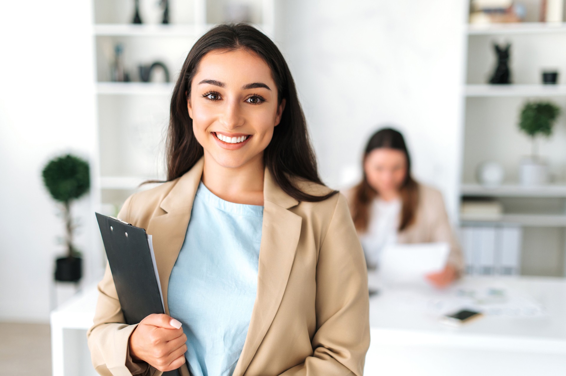 Amazing successful brunette indian or arabian business woman, secretary, company ceo, in a elegant suit stand in a modern office, holding folder with documents, looking and smiles at camera